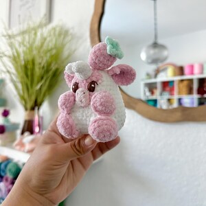 May include: A pink and white crocheted stuffed animal shaped like a cow with a green flower on its head. The cow is being held in a person's hand.