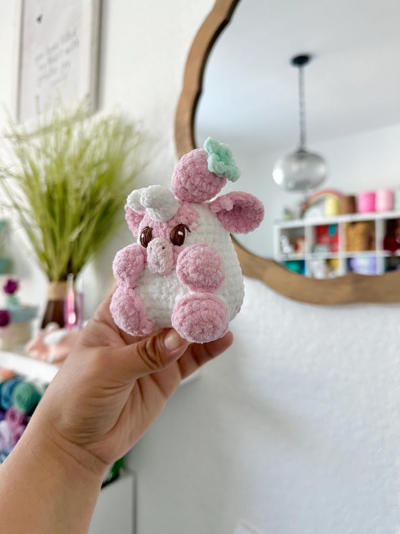 May include: A pink and white crocheted stuffed animal shaped like a cow with a green flower on its head. The cow is being held in a person's hand.