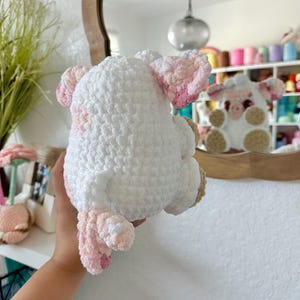 May include: A white crocheted sheep plush toy with pink accents. The sheep is holding a pink flower in its mouth.