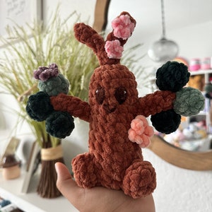 Pode incluir: Um brinquedo de árvore de crochê marrom com flores de crochê verdes e rosa. A árvore tem um rosto sorridente e está sendo segurada na mão de uma pessoa.