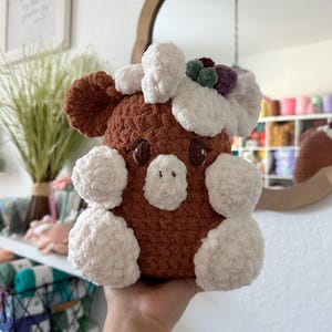 May include: A brown and white crocheted stuffed animal with a flower crown. The animal has a rounded body with white accents and a brown face with black eyes.