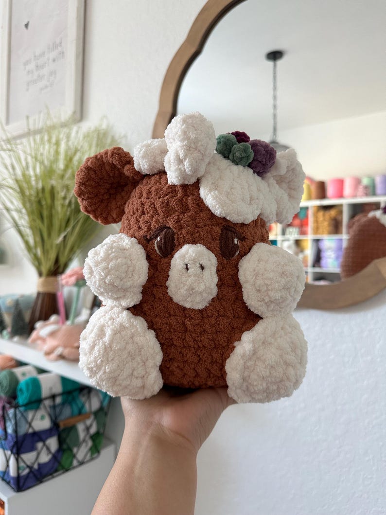 May include: A brown and white crocheted stuffed animal with a flower crown. The animal has a rounded body with white accents and a brown face with black eyes.