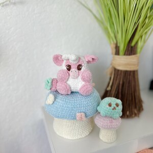 May include: A pink crocheted pig sitting on a blue and white crocheted mushroom. A smaller green crocheted mushroom is next to the larger mushroom.