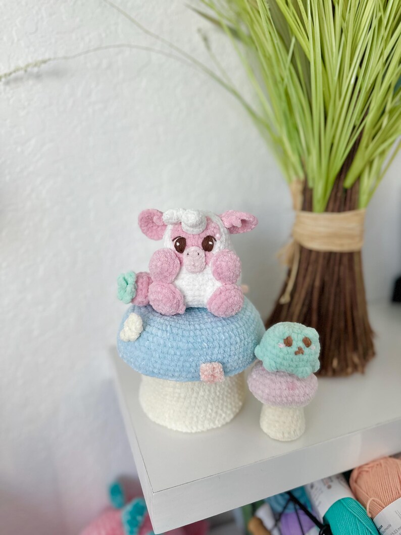 May include: A pink crocheted pig sitting on a blue and white crocheted mushroom. A smaller green crocheted mushroom is next to the larger mushroom.