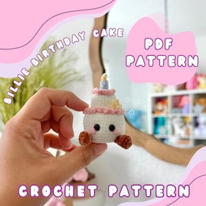 May include: A crocheted birthday cake with a candle, held in a hand. The cake is white with pink and yellow accents. Text on the image reads "BILLIE BIRTHDAY CAKE" and "CROCHET PATTERN". A pink bubble reads "PDF PATTERN".