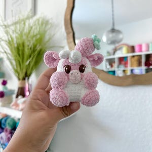 May include: A pink and white crocheted stuffed toy with a green flower on its head. The toy has large eyes and a small nose.