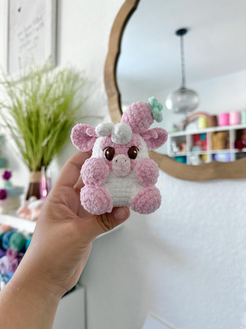 May include: A pink and white crocheted stuffed toy with a green flower on its head. The toy has large eyes and a small nose.