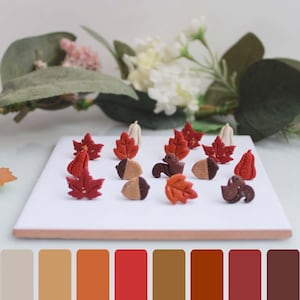 Könnte beinhalten: Eine Sammlung herbstlicher Miniaturdekorationen, darunter rote und orangefarbene Ahornblätter, Eicheln und kleine Kürbisse, angeordnet auf einem weißen Quadrat. Die Farbpalette weist warme Töne von Rot, Orange und Braun auf und erinnert an eine Herbsternte.