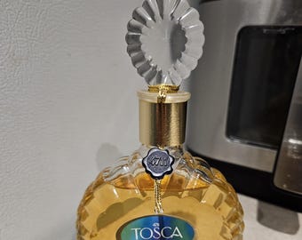 Puede incluir: Una botella de vidrio transparente de Eau de Cologne Tosca con una tapa dorada y un tapón decorativo en forma de flor. La botella tiene una superficie texturizada y una etiqueta con el nombre de la marca y la descripción del producto.