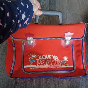 May include: Red canvas satchel with a black handle and two silver clasps. The bag has a blue and yellow trim and features a cartoon image of two children with the text "Love Ya Patches" and "The World of Patches McGee © Gabriel Industries, Inc."