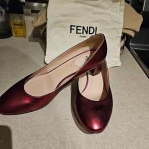 Puede incluir: Un par de zapatos de tacón color burdeos metalizado con punta redondeada y tacón bajo cuadrado. El forro interior es de color beige claro. Una bolsa de tela blanca con el texto negro FENDI ROMA se sitúa detrás de los zapatos.