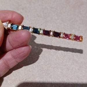 以下が含まれることがあります： カラフルな宝石が並んだ金のブレスレット。宝石は金の枠にセットされ、小さなダイヤモンドで囲まれています。ブレスレットは美しくエレガントなジュエリーです。
