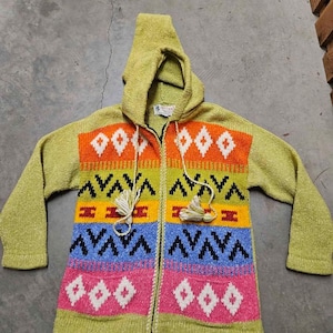 May include: A lime green knit zip-up hoodie featuring a multi-coloured horizontal pattern with orange, yellow, blue, and pink stripes. The garment includes black geometric accents, white diamond shapes, and two drawstrings with tassels at the ends.