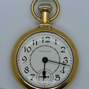 May include: A gold-toned pocket watch with a white face and black hands. The watch has Roman numeral hour markers and a small second hand dial. The watch face also has the text "WESTCLOX" printed on it.