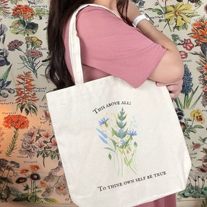 Pode incluir: Uma bolsa tote de lona branca com um design floral e o texto "This above all: To thine own self be true".