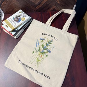 Pode incluir: Uma bolsa tote de lona bege com al&ccedil;as pretas e um design floral. A bolsa tem o texto "This above all: To thine own self be true" impresso nela.