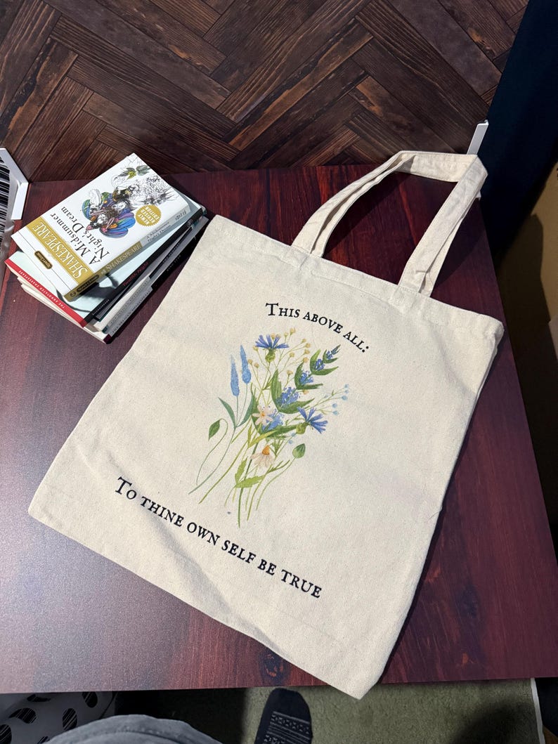 Pode incluir: Uma bolsa tote de lona bege com al&ccedil;as pretas e um design floral. A bolsa tem o texto "This above all: To thine own self be true" impresso nela.