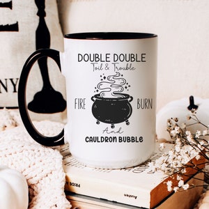 May include: White ceramic mug with a black rim and handle. The mug features a black and white illustration of a cauldron with steam rising from it. The text "DOUBLE DOUBLE Toil & Trouble FIRE BURN And CAULDRON BUBBLE" is printed on the mug.