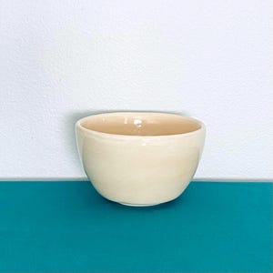 May include: A small, cream-colored ceramic bowl with a rounded shape. The bowl has a smooth, glossy finish and is set against a white and teal background. The bowl is likely used for serving food or as a decorative item.