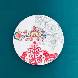 Pu&ograve; includere: Piatto in ceramica bianca con un colorato motivo floreale e motivi di cavalli rossi. Il piatto presenta un motivo floreale dettagliato nelle tonalit&agrave; del blu, giallo e rosso, con un motivo geometrico grigio sul lato destro. Il piatto &egrave; posto su uno sfondo verde acqua.