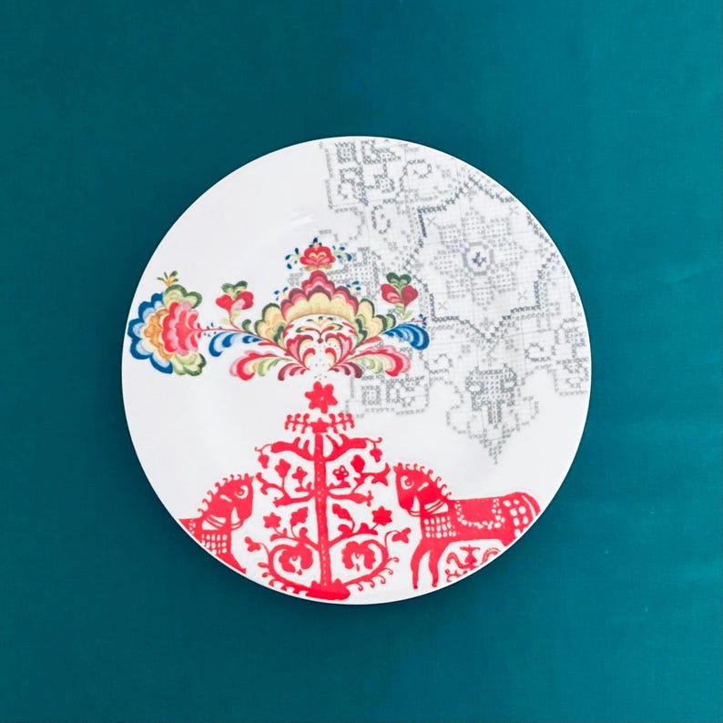 Pu&ograve; includere: Piatto in ceramica bianca con un colorato motivo floreale e motivi di cavalli rossi. Il piatto presenta un motivo floreale dettagliato nelle tonalit&agrave; del blu, giallo e rosso, con un motivo geometrico grigio sul lato destro. Il piatto &egrave; posto su uno sfondo verde acqua.