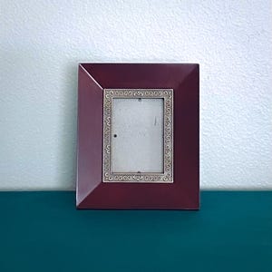 May include: A rectangular picture frame with a wide, smooth, deep burgundy border. The inner section features a metallic silver-toned trim decorated with a repeating embossed scrollwork pattern surrounding the central rectangular opening.
