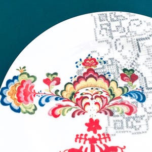 Pu&ograve; includere: Un piatto in ceramica bianca con un colorato motivo floreale. Il design presenta fiori e foglie rosse, gialle, blu e verdi. Sullo sfondo &egrave; visibile un motivo a punto croce grigio.