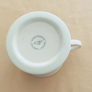 May include: White ceramic mug with a handle, seen from below. The base features a circular emblem with a bird illustration and text. The mug is labelled "Microwave & Dishwasher Safe" and "Made in China".