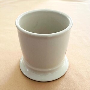 May include: A white ceramic shaving mug. The mug has a wide base and a cylindrical body. It has a smooth, glossy finish and a slightly flared rim. The mug is empty and appears unused.