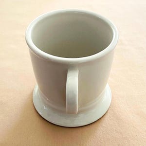 May include: A cream-coloured ceramic mug with a handle and a wide base. The mug has a simple design and appears to be empty. The mug is on a light brown surface.