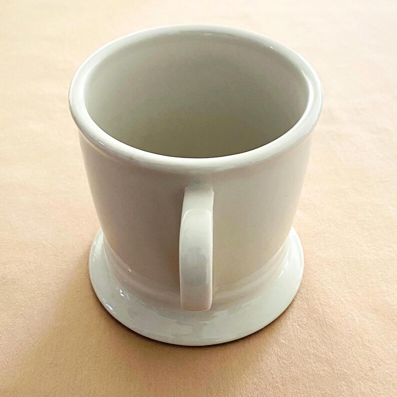 May include: A cream-coloured ceramic mug with a handle and a wide base. The mug has a simple design and appears to be empty. The mug is on a light brown surface.
