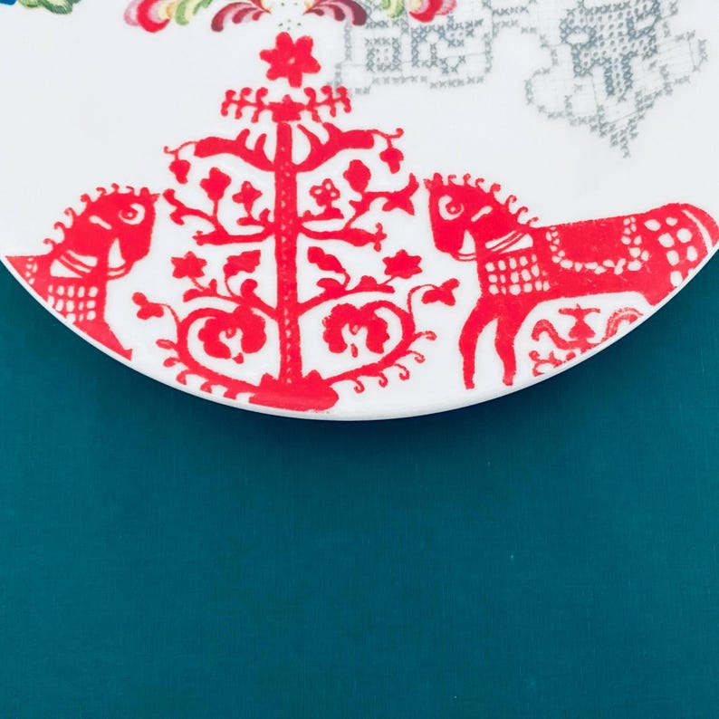 Pu&ograve; includere: Un piatto in ceramica bianca con un design di arte popolare rosso. Il design include un motivo ad albero centrale e due figure di cavallo a dondolo. Il piatto ha un bordo curvo ed &egrave; posto su uno sfondo verde acqua.