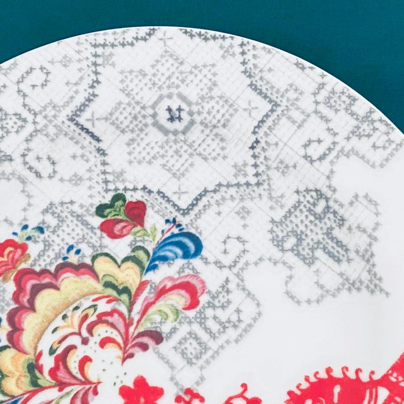 Pu&ograve; includere: Un piatto in ceramica bianca con un colorato motivo floreale. Il piatto presenta un motivo dettagliato con elementi floreali rossi, gialli, blu e verdi. Il bordo ha un motivo grigio simile al punto croce.