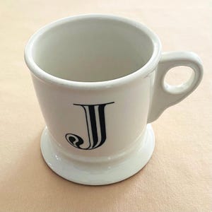 May include: White ceramic mug featuring a black letter "J". The mug has a rounded base and a handle. The mug is on a light tan surface. The mug is likely used for drinking tea or coffee.