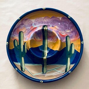 May include: A hand-painted ceramic serving bowl with a Southwestern desert landscape design. The bowl features a blue rim and a central dip area. The interior depicts cacti, mountains, and a sunset in shades of orange, purple, and blue.