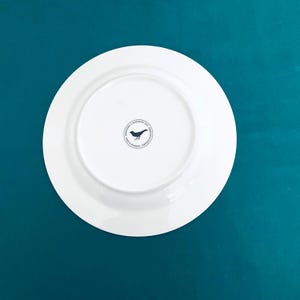 Pu&ograve; includere: Un piatto in ceramica bianca con un bordo largo, visto dal basso. Un logo circolare in bianco e nero con un uccello &egrave; centrato sul piatto. Il piatto &egrave; posto su uno sfondo turchese.