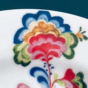 Pu&ograve; includere: Primo piano di un piatto in ceramica bianca con un vivace motivo floreale. Il fiore presenta petali nelle tonalit&agrave; del rosso, rosa, blu, arancione e verde. Il design &egrave; dettagliato e intricato, che mostra un'estetica colorata e dipinta a mano.