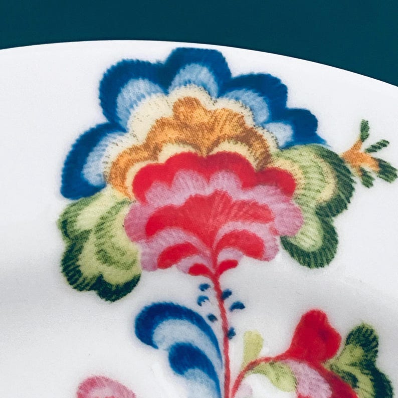Pu&ograve; includere: Primo piano di un piatto in ceramica bianca con un vivace motivo floreale. Il fiore presenta petali nelle tonalit&agrave; del rosso, rosa, blu, arancione e verde. Il design &egrave; dettagliato e intricato, che mostra un'estetica colorata e dipinta a mano.