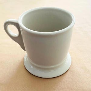 May include: A white ceramic mug with a rounded handle and a pedestal base. The mug has a smooth, glossy finish and a simple, classic design. The mug is approximately 10 cm tall.