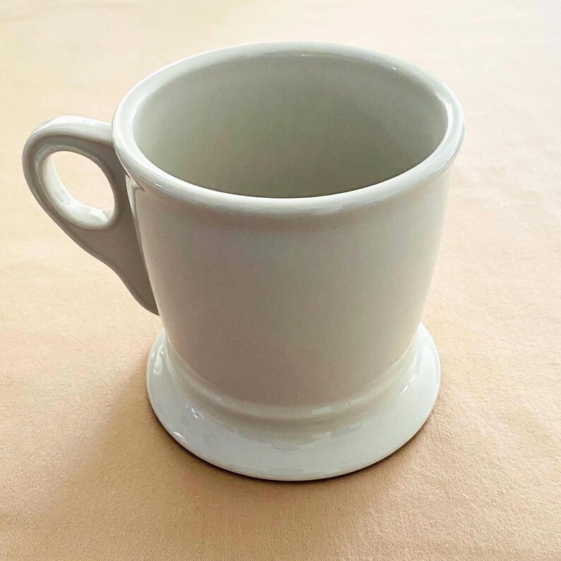 May include: A white ceramic mug with a rounded handle and a pedestal base. The mug has a smooth, glossy finish and a simple, classic design. The mug is approximately 10 cm tall.