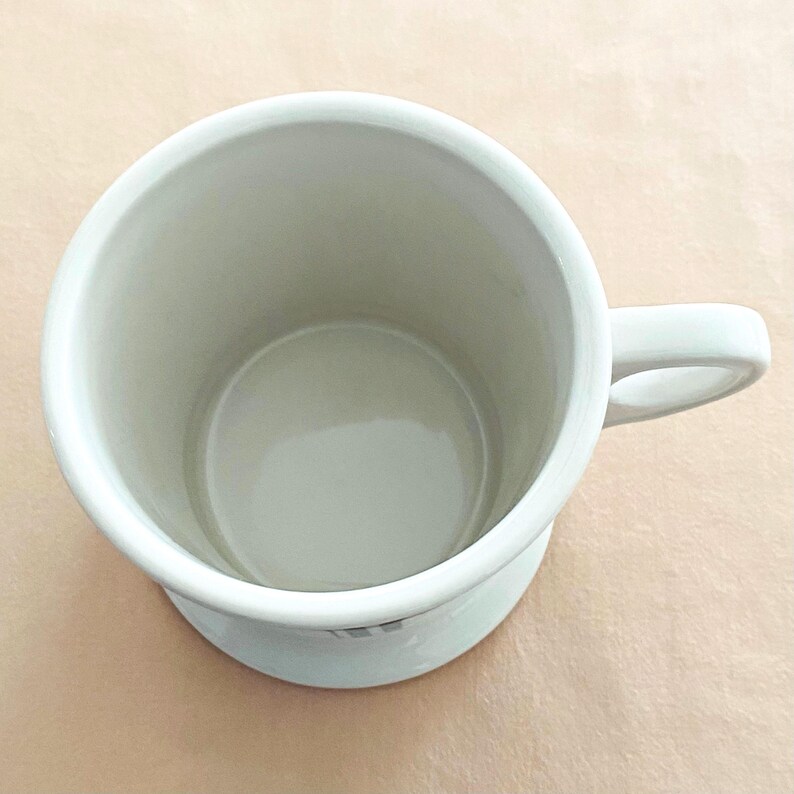 May include: A white ceramic mug with a rounded base and a curved handle. The mug has a smooth, glossy finish and a simple, minimalist design. The interior of the mug is a light grey colour.