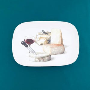 May include: White rectangular serving platter with a watercolor illustration of cheese, a glass of red wine, and a cheese knife. The cheese varieties include a wrapped block, a wedge, and a round cheese wheel.