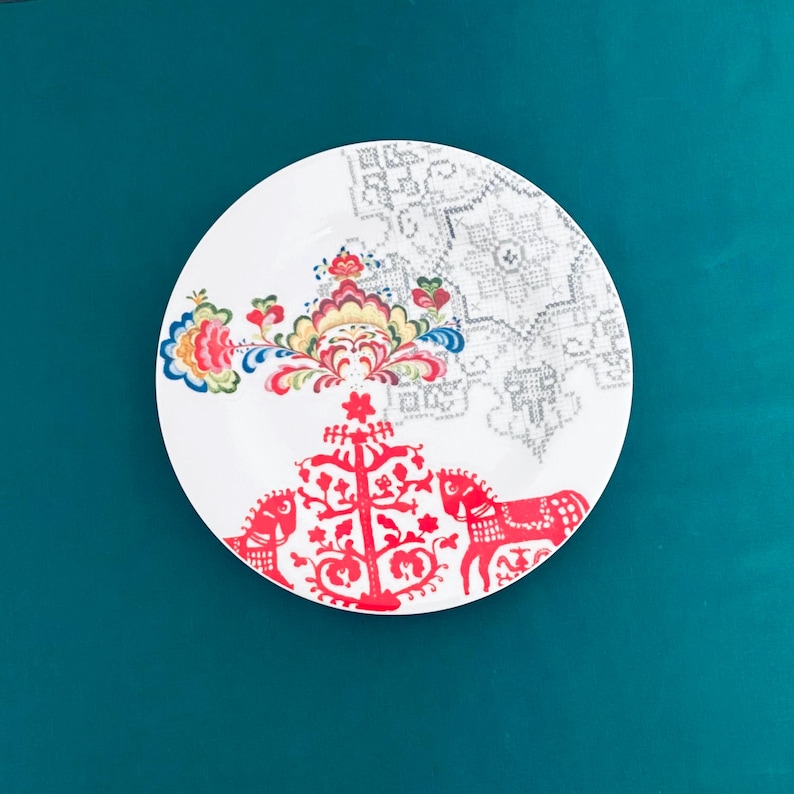 Pu&ograve; includere: Piatto in ceramica bianca con un colorato motivo floreale e motivi di cavalli rossi. Il piatto presenta un motivo dettagliato in rosso, blu, giallo e verde, con un disegno geometrico grigio sul lato superiore destro. Il piatto &egrave; posto su uno sfondo verde acqua.