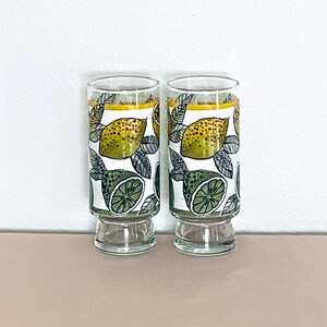 May include: Two clear glass tumblers featuring a repeating pattern of yellow lemons and green lime halves with black outlined leaves. Each glass has a thick, weighted base and a thin yellow horizontal band near the top rim of the design.