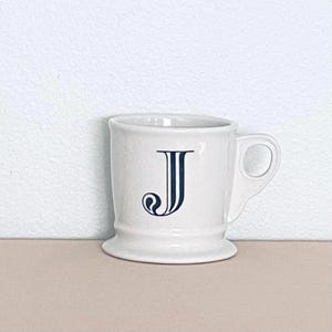 May include: A white ceramic mug with a handle, displaying a navy blue letter "J" in an ornate style. The mug has a flared base and a classic design, ideal for tea or coffee. The mug is a standard size.