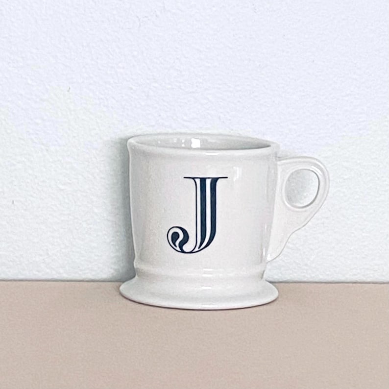 May include: A white ceramic mug with a handle, displaying a navy blue letter "J" in an ornate style. The mug has a flared base and a classic design, ideal for tea or coffee. The mug is a standard size.