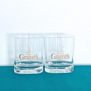 May include: Two clear, lowball whisky glasses. Each glass has the words "William Grant's" in gold lettering, with a crest design above the name. The glasses are on a teal surface against a white background.