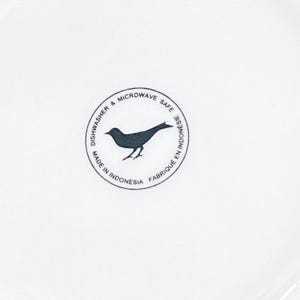 Pu&ograve; includere: Un piatto in ceramica bianco con un logo di uccello nero al centro. Il logo &egrave; all'interno di un bordo circolare con le parole "Dishwasher & Microwave Safe" e "Made in Indonesia".