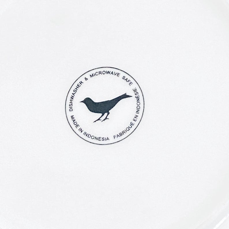 Pu&ograve; includere: Un piatto in ceramica bianco con un logo di uccello nero al centro. Il logo &egrave; all'interno di un bordo circolare con le parole "Dishwasher & Microwave Safe" e "Made in Indonesia".