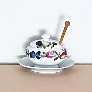 May include: A white ceramic lidded jar with a matching saucer and a wooden honey dipper. The jar and lid feature a colorful botanical pattern of blue flowers, pink roses, and pears with green leaves. The lid has a small knob handle.